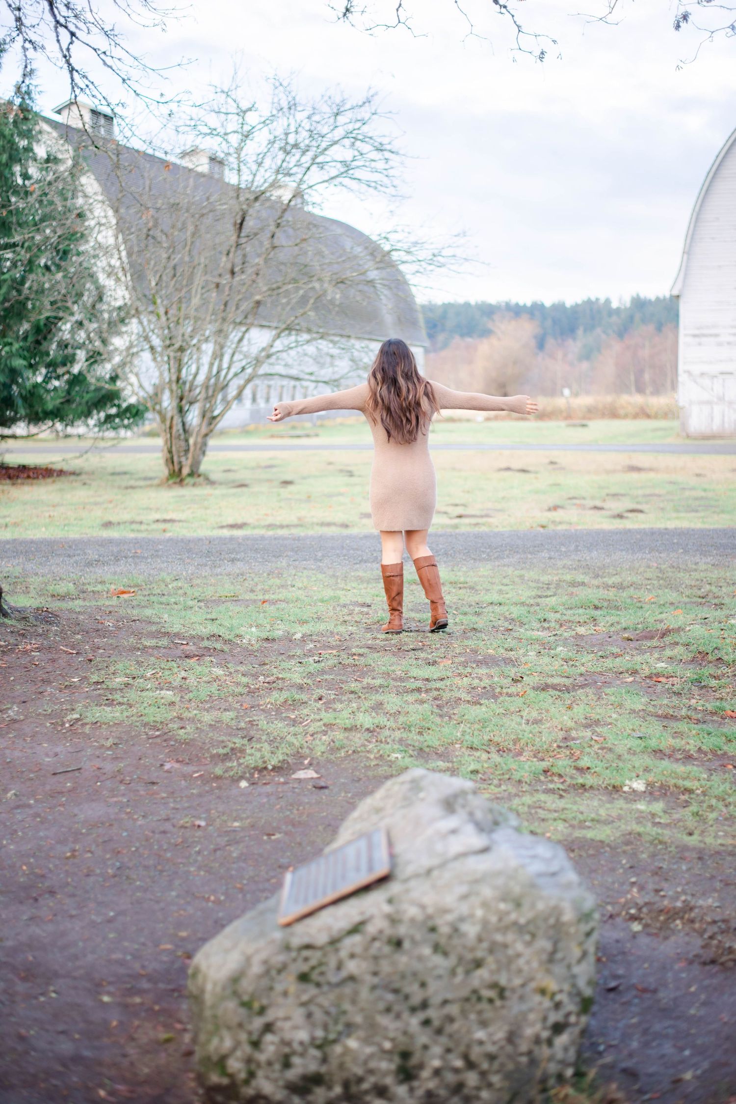 Person in beige sweater dress and brown boots standing in a rural farm setting with white barns.