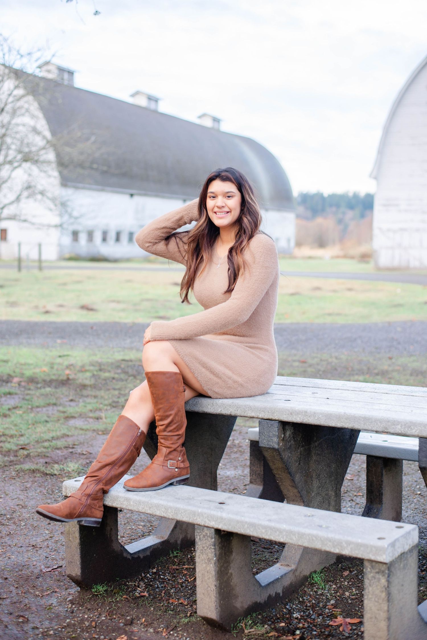 Woman in beige dress and knee-high boots sitting on wooden bench with white barn in background.