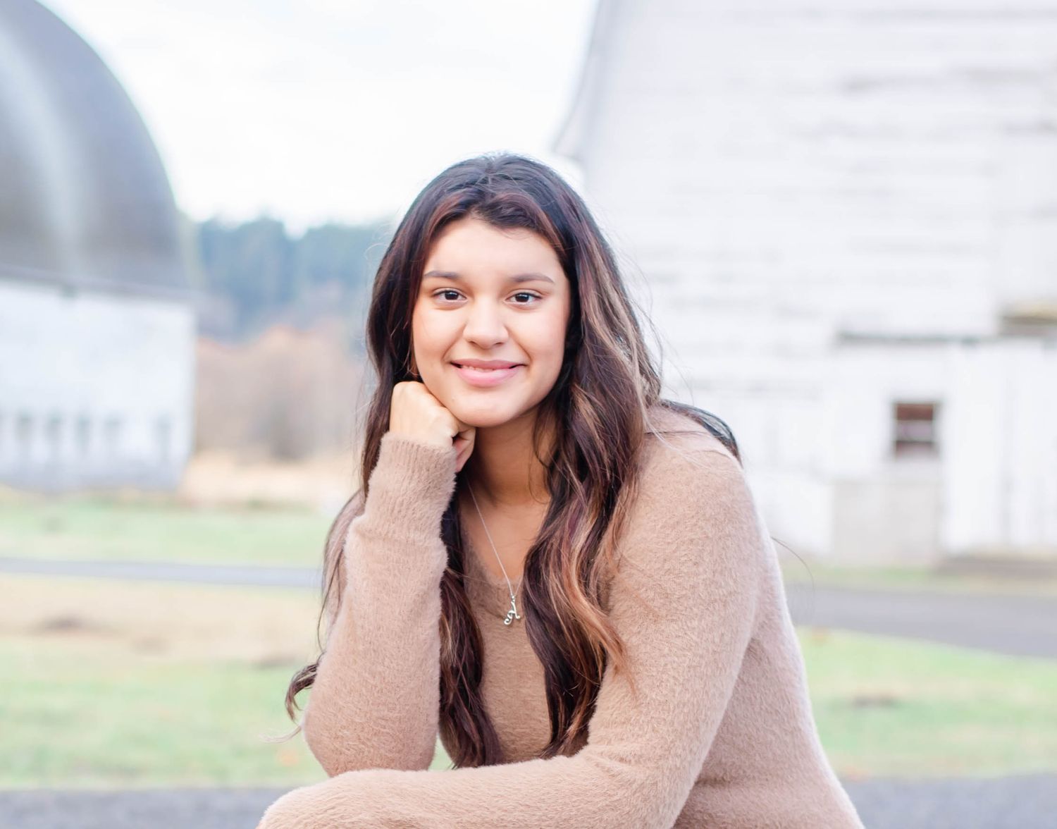 Portrait of woman in beige sweater with long dark hair against rural farm backdrop.