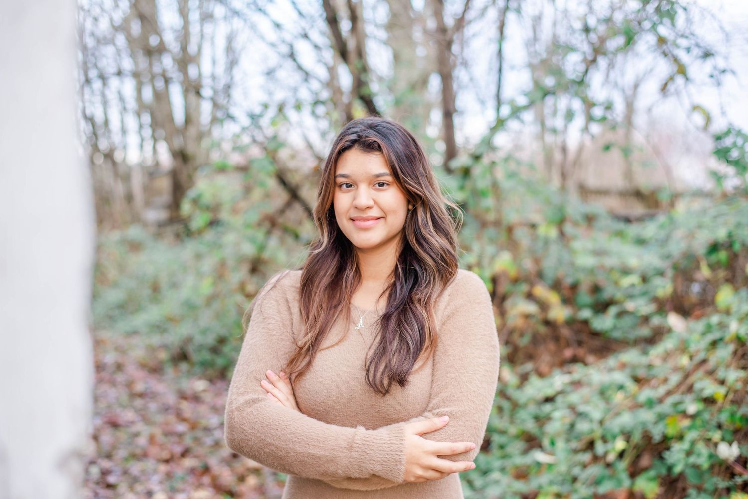 Woman in beige sweater smiling with scenic winter foliage in background.