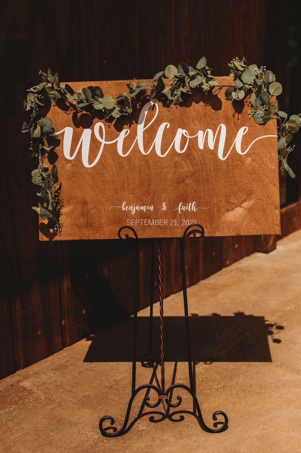 Wooden welcome sign decorated with greenery garland on metal stand at wedding venue entrance.