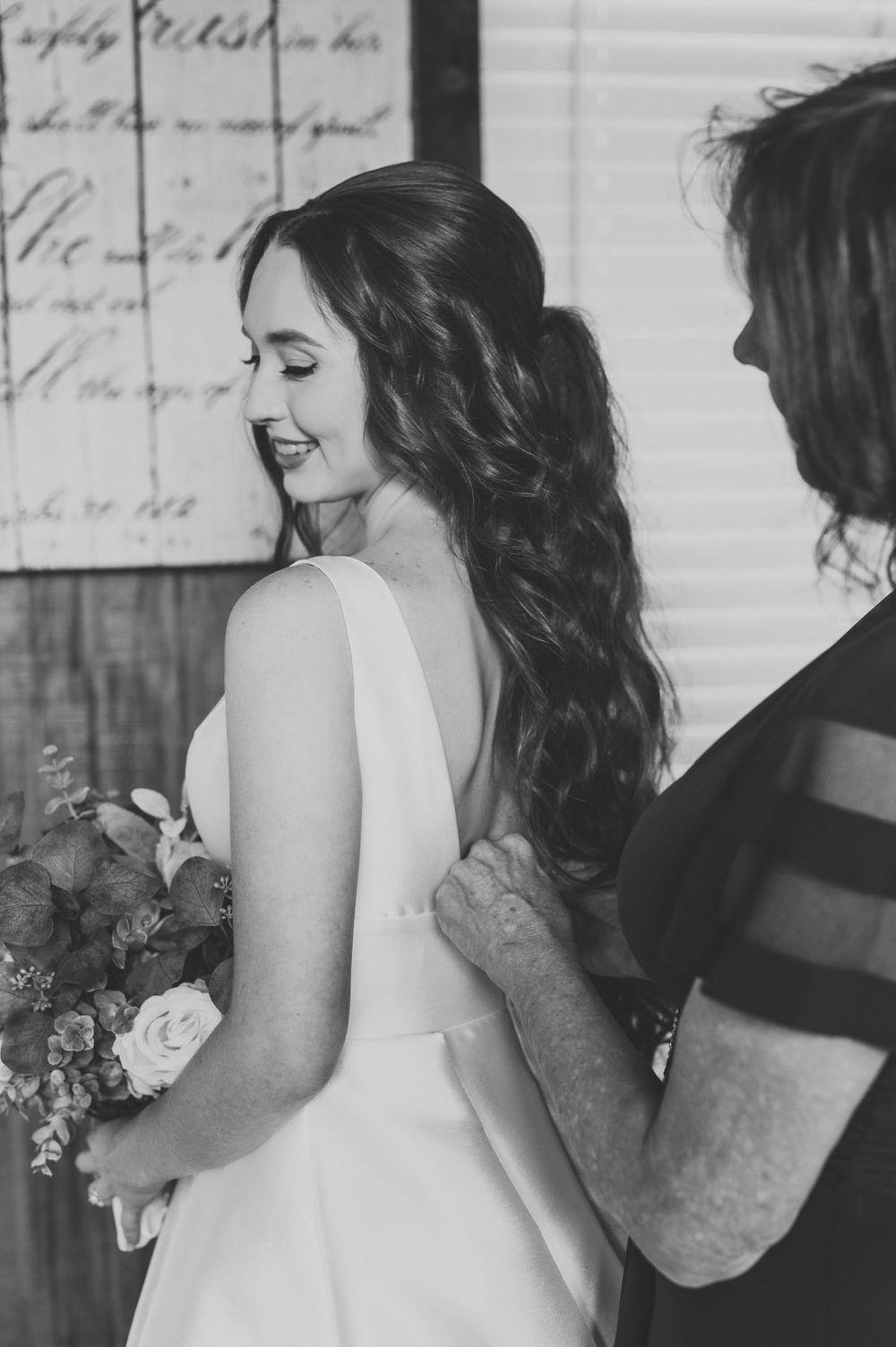 Black and white photo of bride being helped with wedding dress preparations.