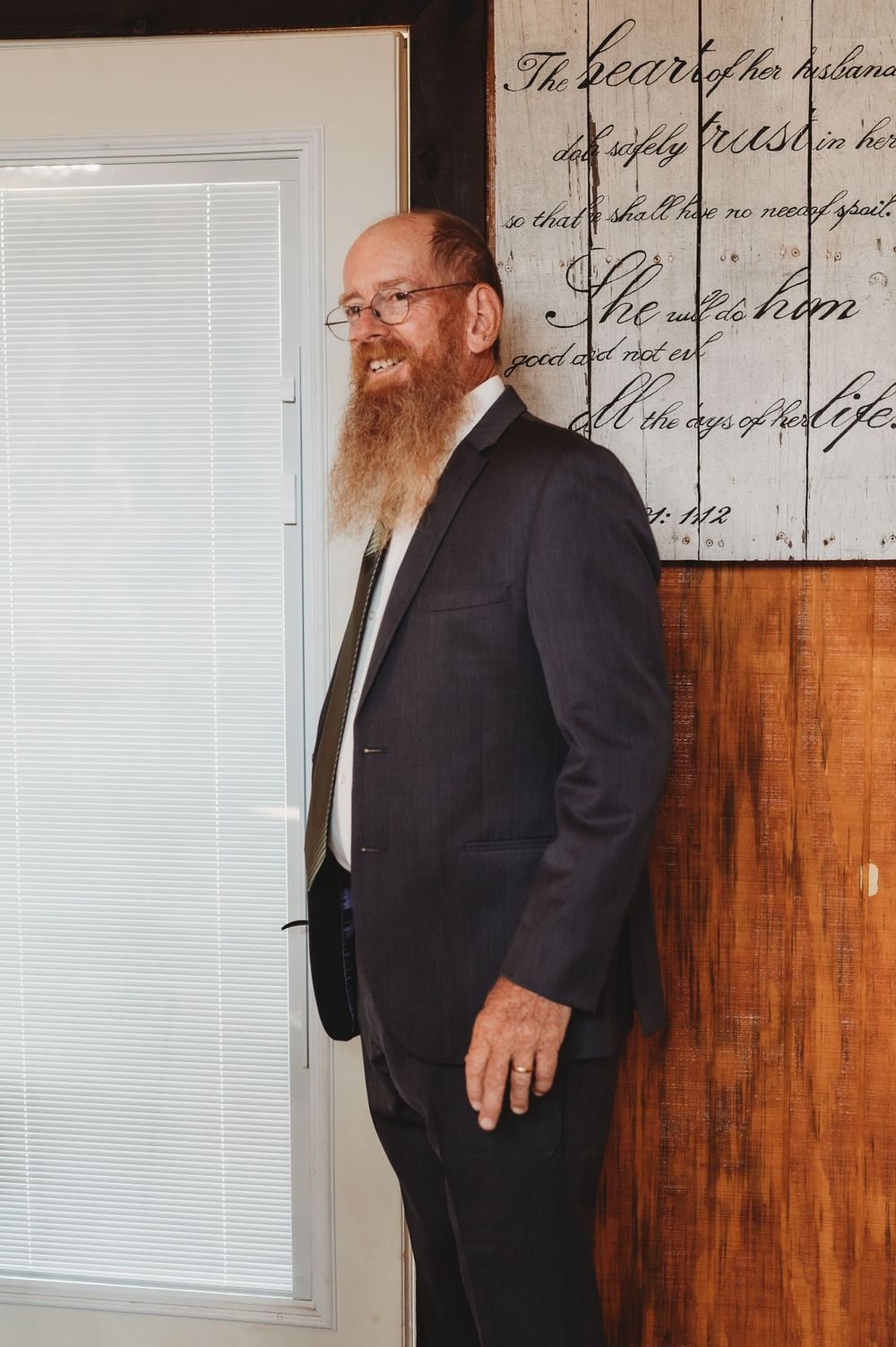 Man in dark suit standing against wooden wall with text artwork.