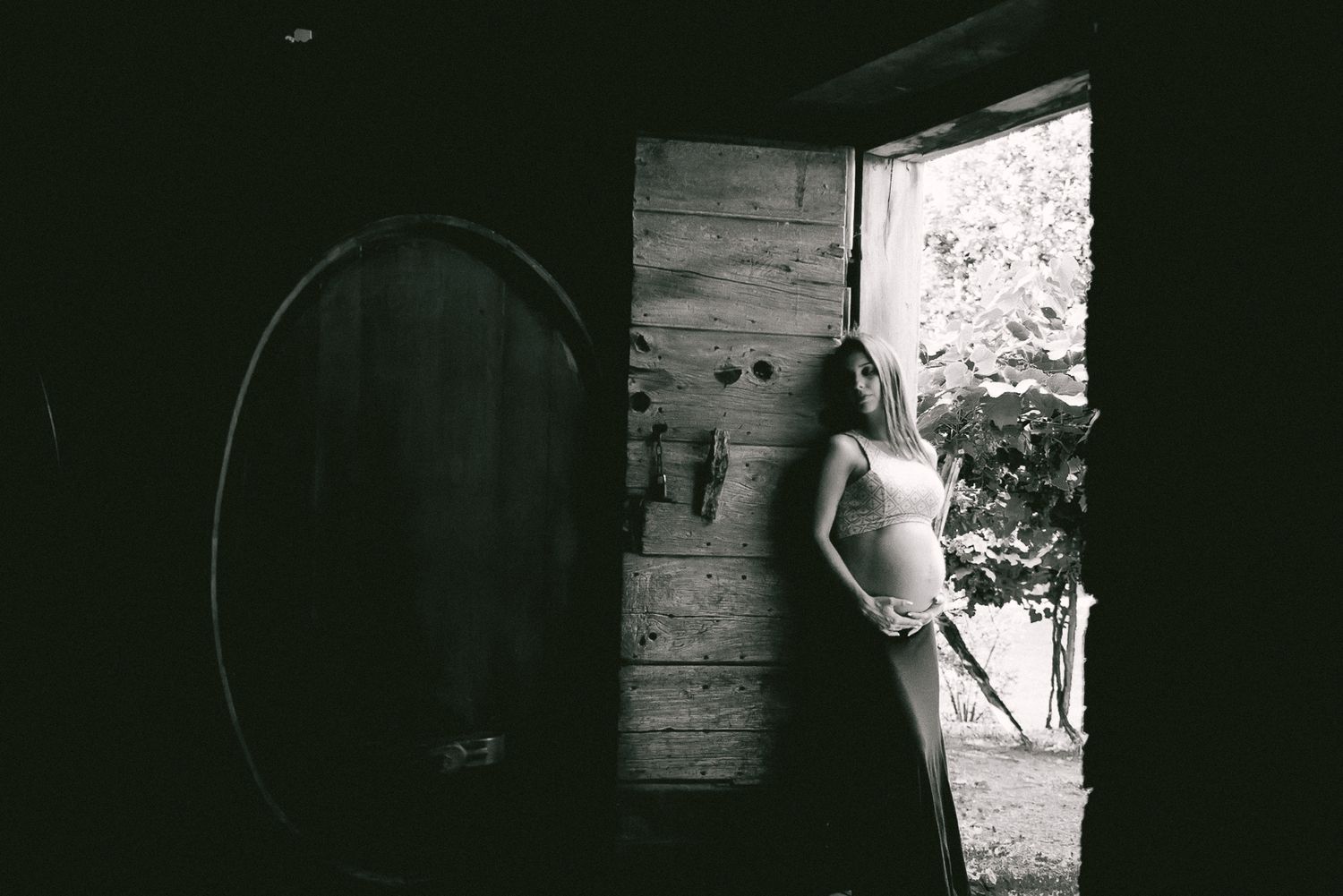 A atmospheric black and white maternity portrait with dramatic side lighting through a doorway.