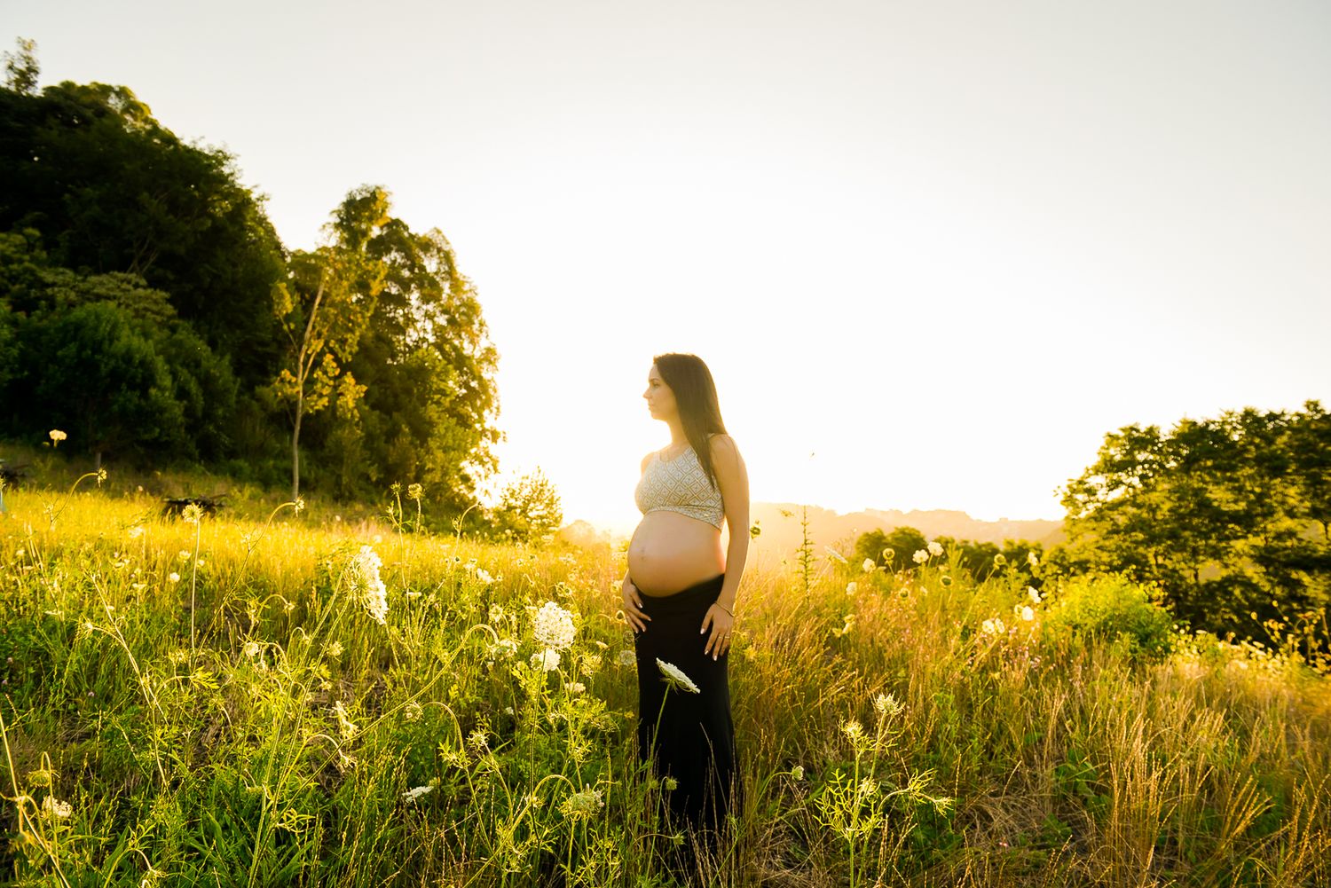 Pregnant figure standing in a sunlit meadow filled with wildflowers during golden hour.