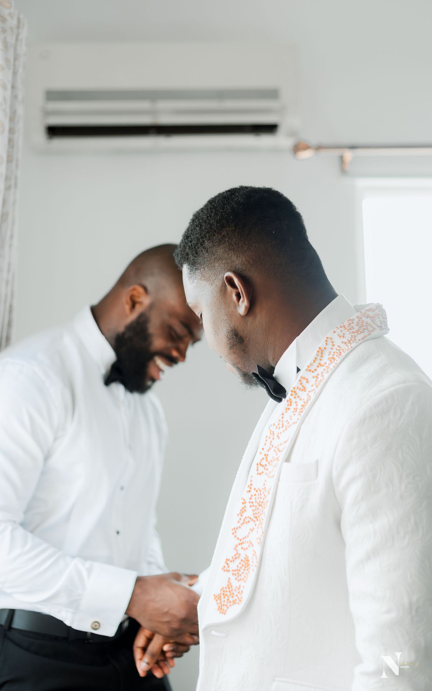 Two men in white formal attire sharing a friendly moment before an event.