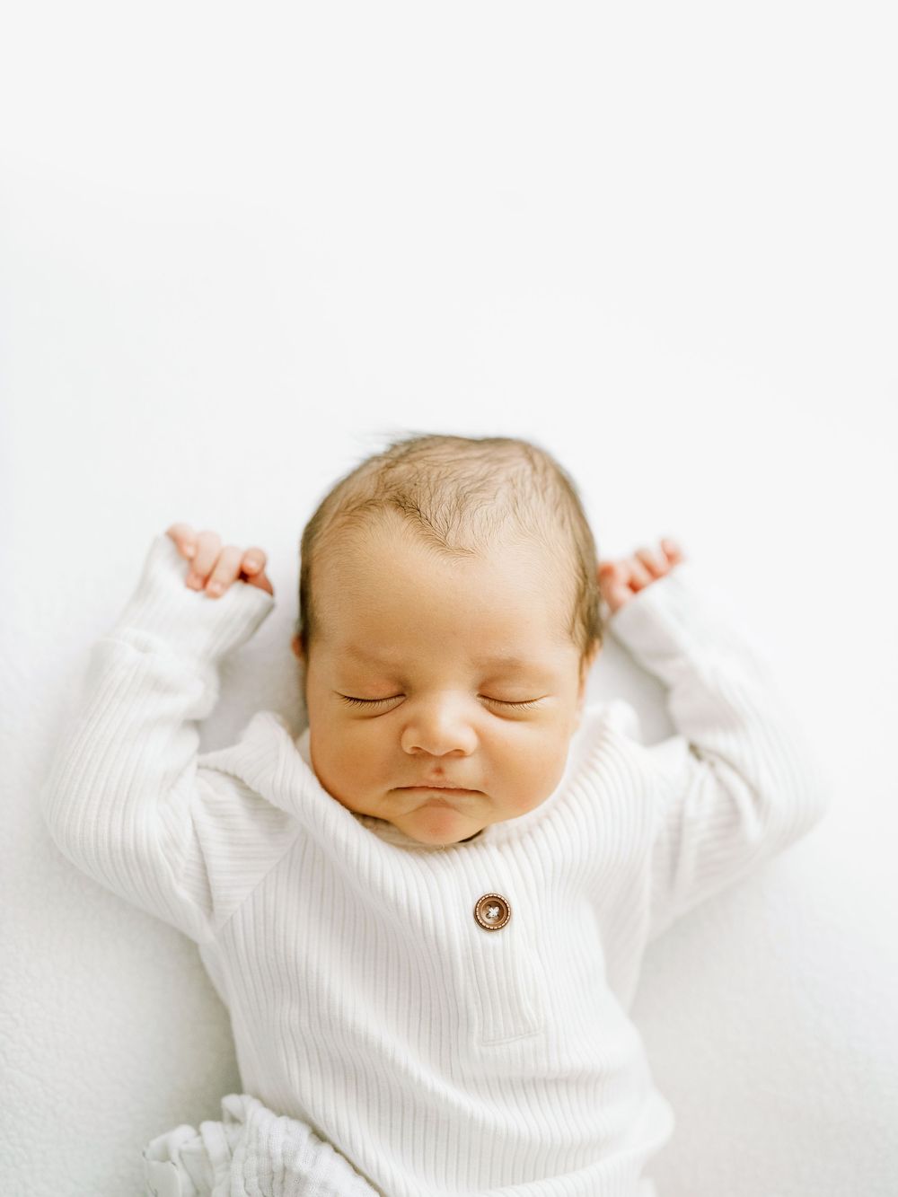 Peaceful sleeping newborn baby wearing white ribbed sweater lies on white background with arms raised.