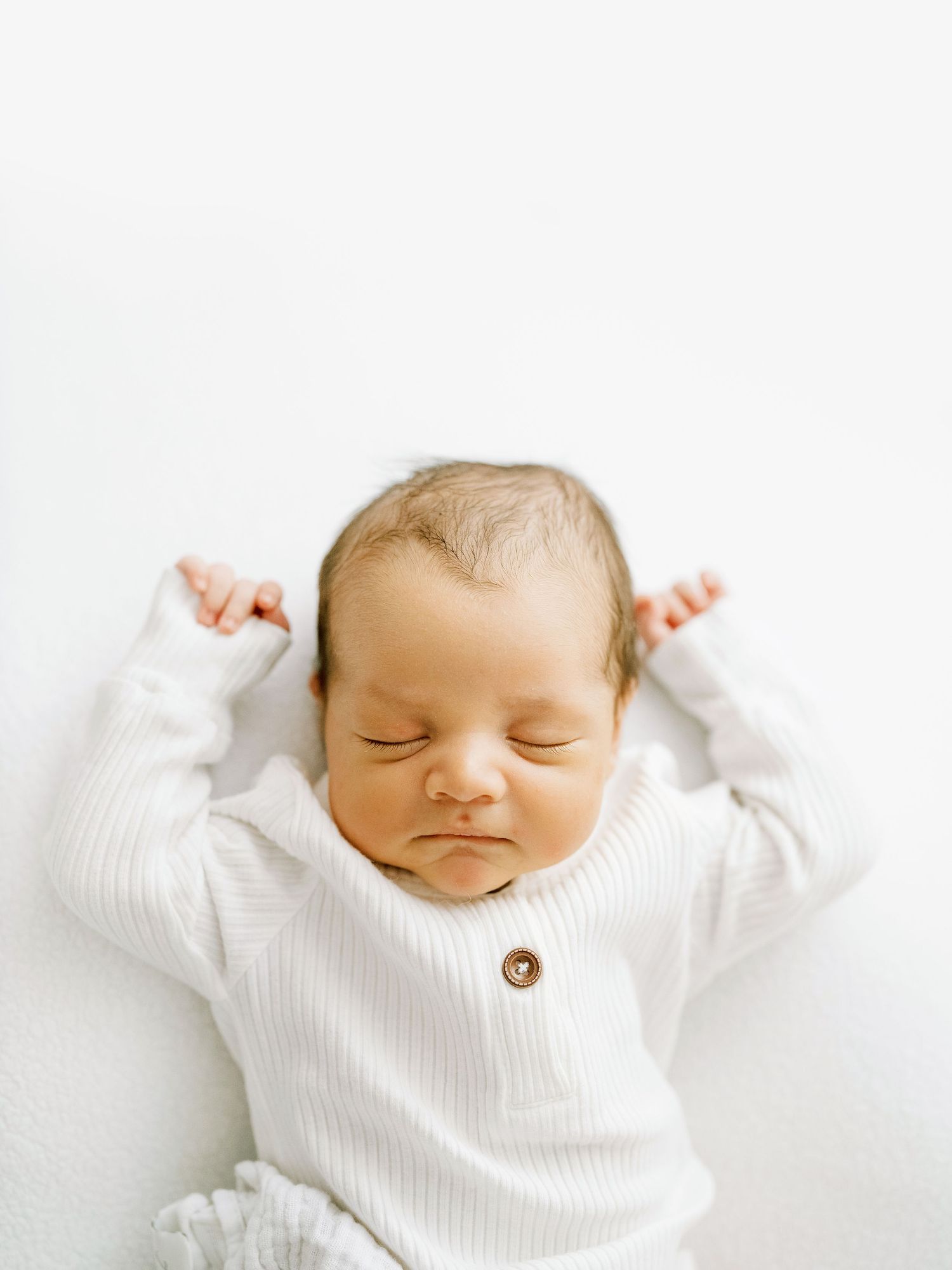 Peaceful sleeping newborn baby wearing white ribbed sweater lies on white background with arms raised.