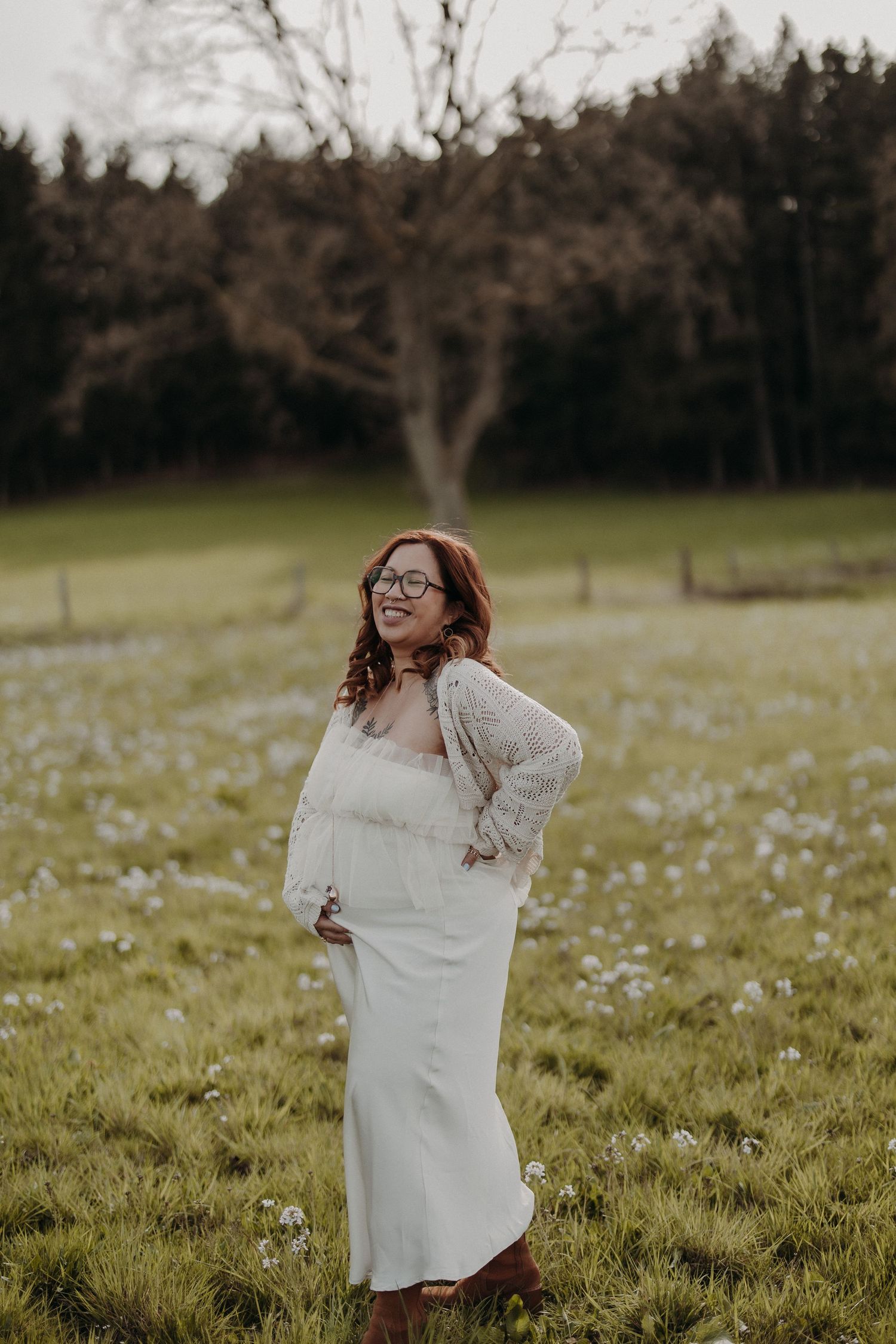 A pregnant woman in glasses laughs joyfully while standing in a field of spring flowers.