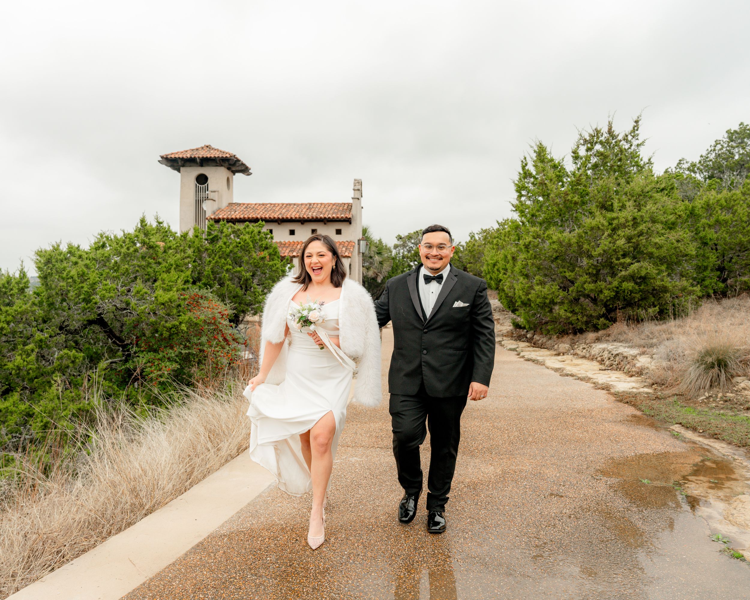 A newly married couple walks together on a rustic dirt path surrounded by green coastal vegetation on their wedding day.