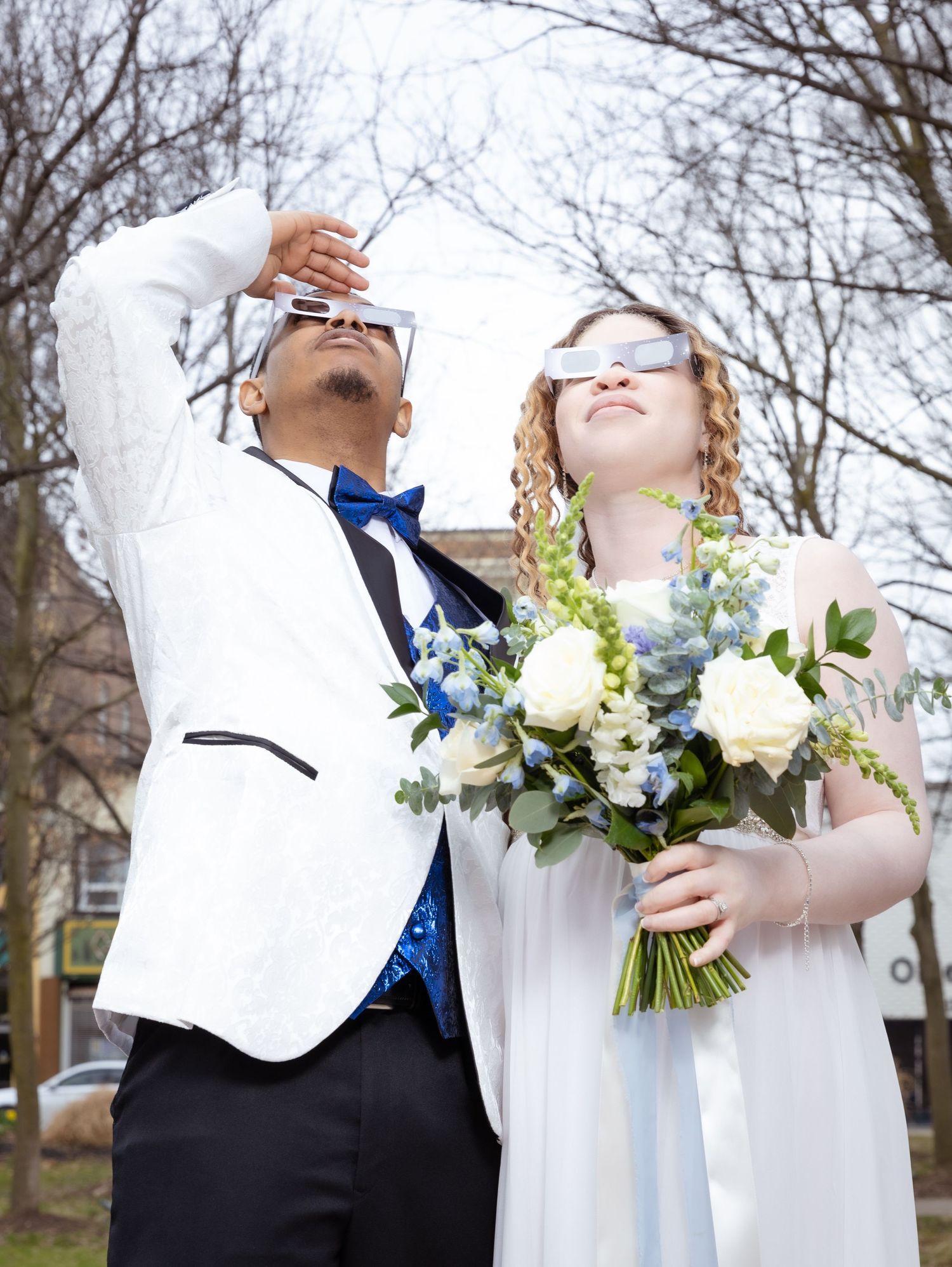 A playful wedding couple poses outdoors with a white and blue floral bouquet against bare winter trees.