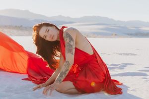 A tattooed figure in a red dress poses elegantly on white sand during golden hour.