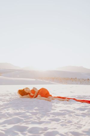 A figure in red reclines on pristine white sand with flowing fabric against desert mountains.