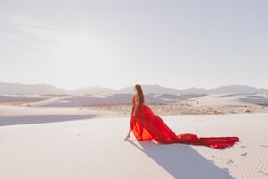 A figure in a flowing red dress poses gracefully against white sand dunes and distant mountains.