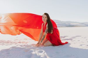 A figure in a flowing red dress poses gracefully on white sand dunes with a billowing orange fabric in the sunlight.