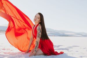 A red-dressed figure sits on white sand as vibrant orange fabric billows dramatically against a desert landscape.