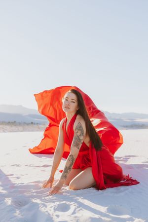 A figure in red kneels gracefully on white sand as orange fabric flows in the desert breeze.