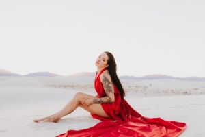 A flowing red dress creates dramatic contrast against white sand dunes and distant mountains in an artistic photo shoot.