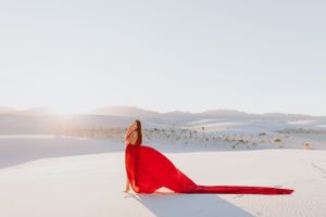A red-dressed figure stands on white sand dunes with flowing fabric against desert mountains.