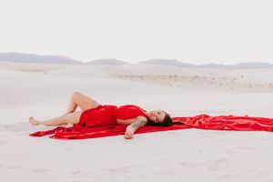 Dramatic pose with red fabric stretching across white sand dunes in minimalist desert landscape.