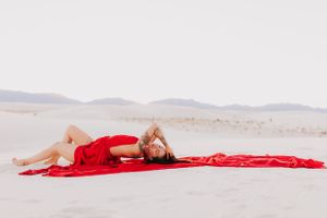 Red dress creates striking visual on white sand with mountains in soft focus background.
