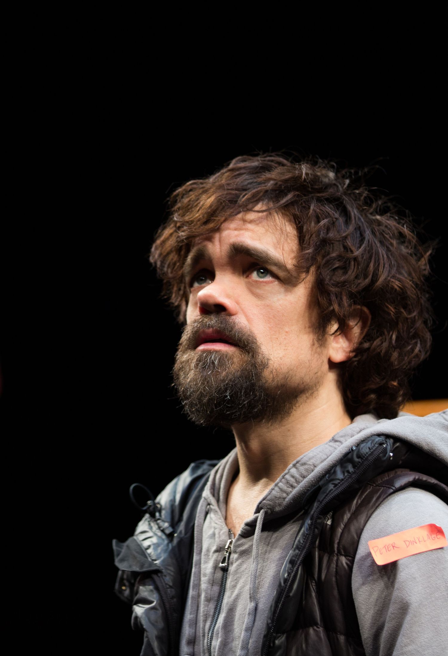 Peter Dinklage rehearsing for a theater performance, shown in profile wearing a denim jacket under dramatic stage lighting.