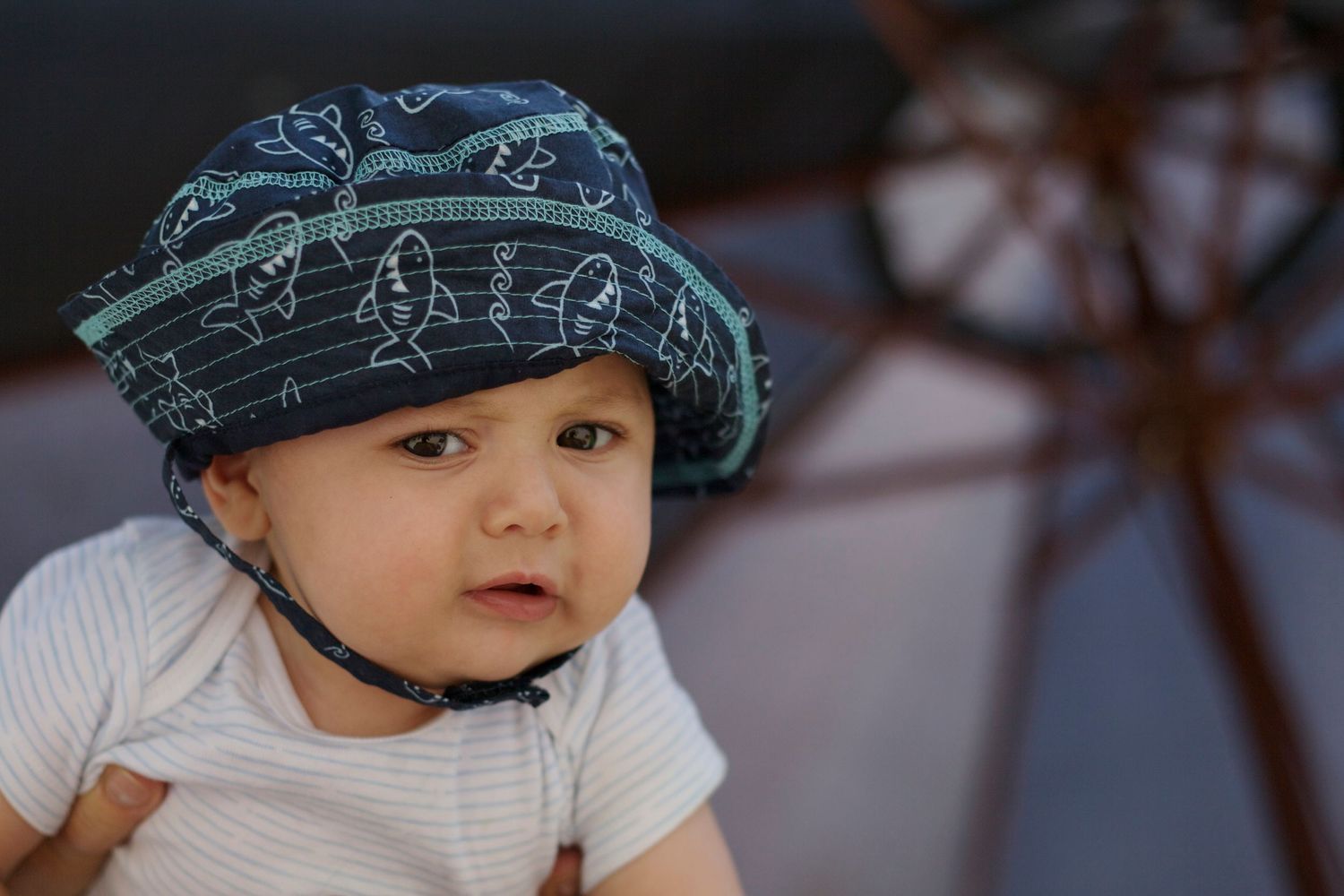 Documentary portrait of six-month-old baby in patterned blue cap and white onesie, showing playful expression at home.