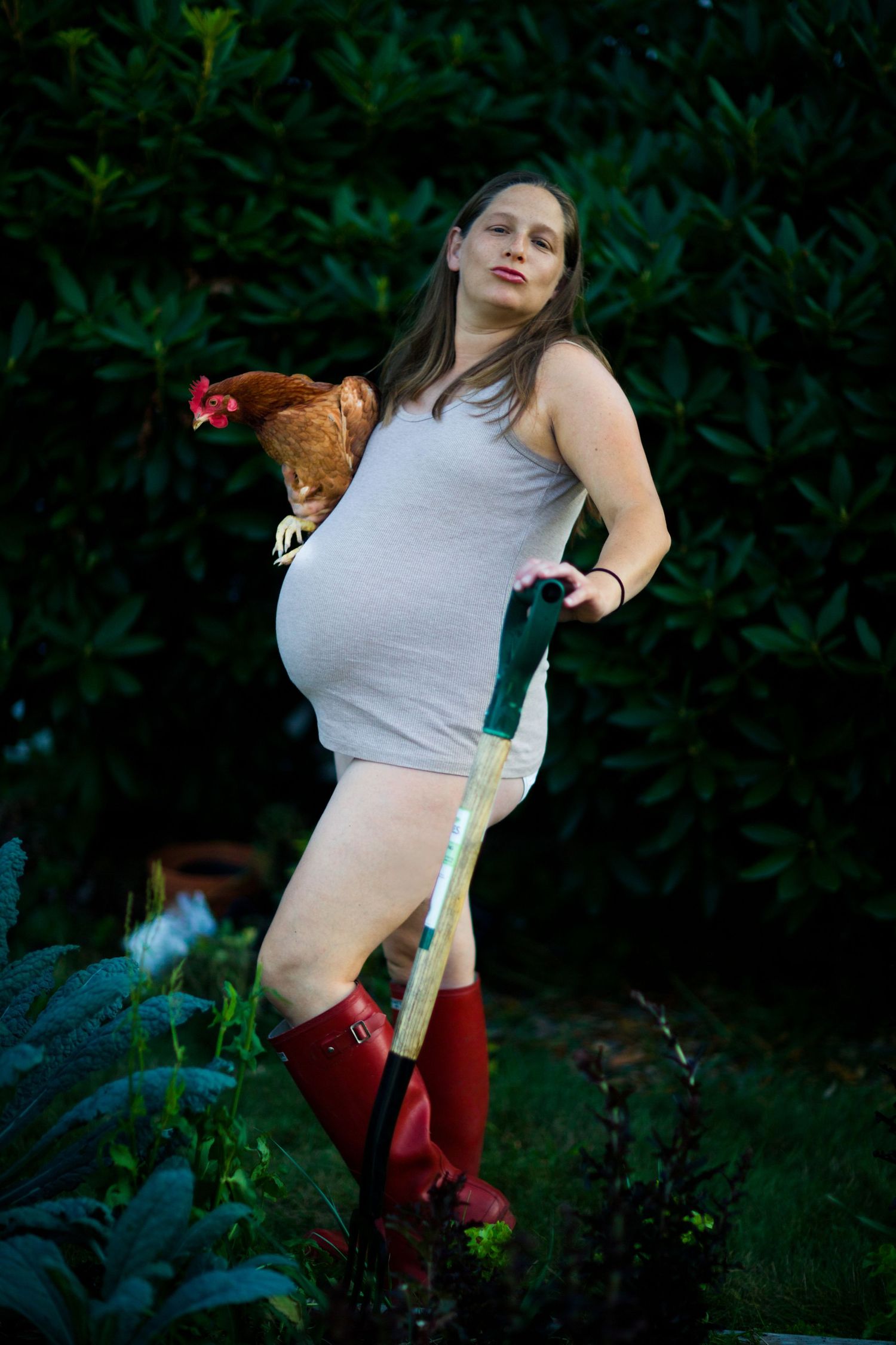 Pregnant woman in muck boots holding chicken and pitchfork in garden, humorous editorial maternity portrait.