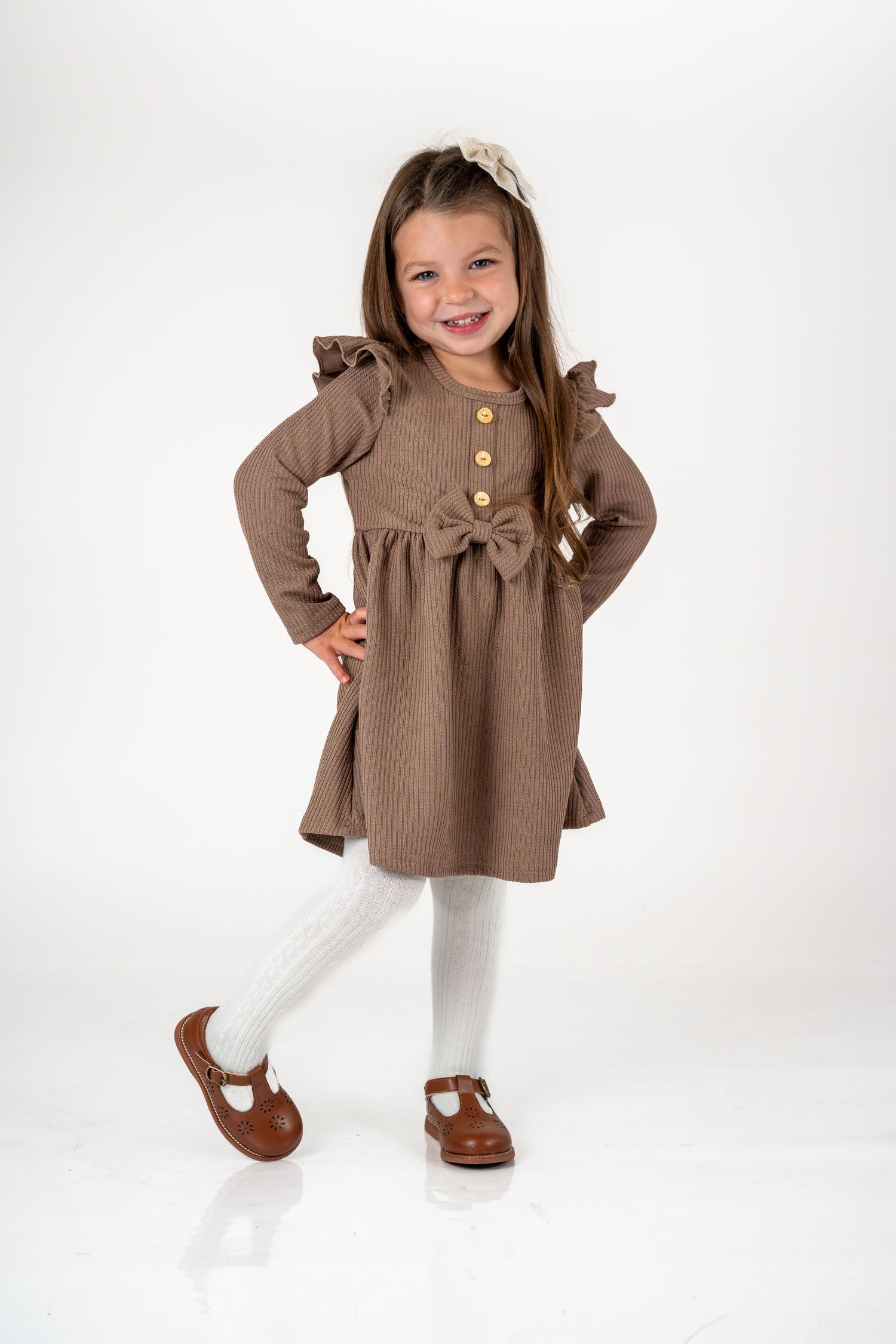 Young model wearing a brown ruffle dress with white tights and brown mary jane shoes against white background.