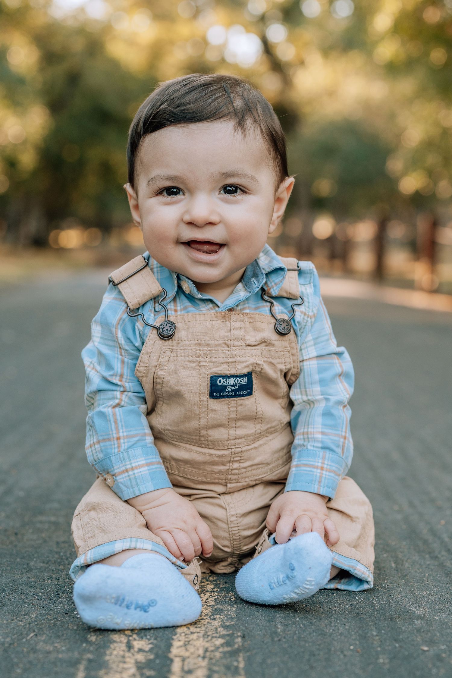 Small child wearing tan overalls and plaid blue shirt sits outdoors with a cheerful expression on an autumn day.