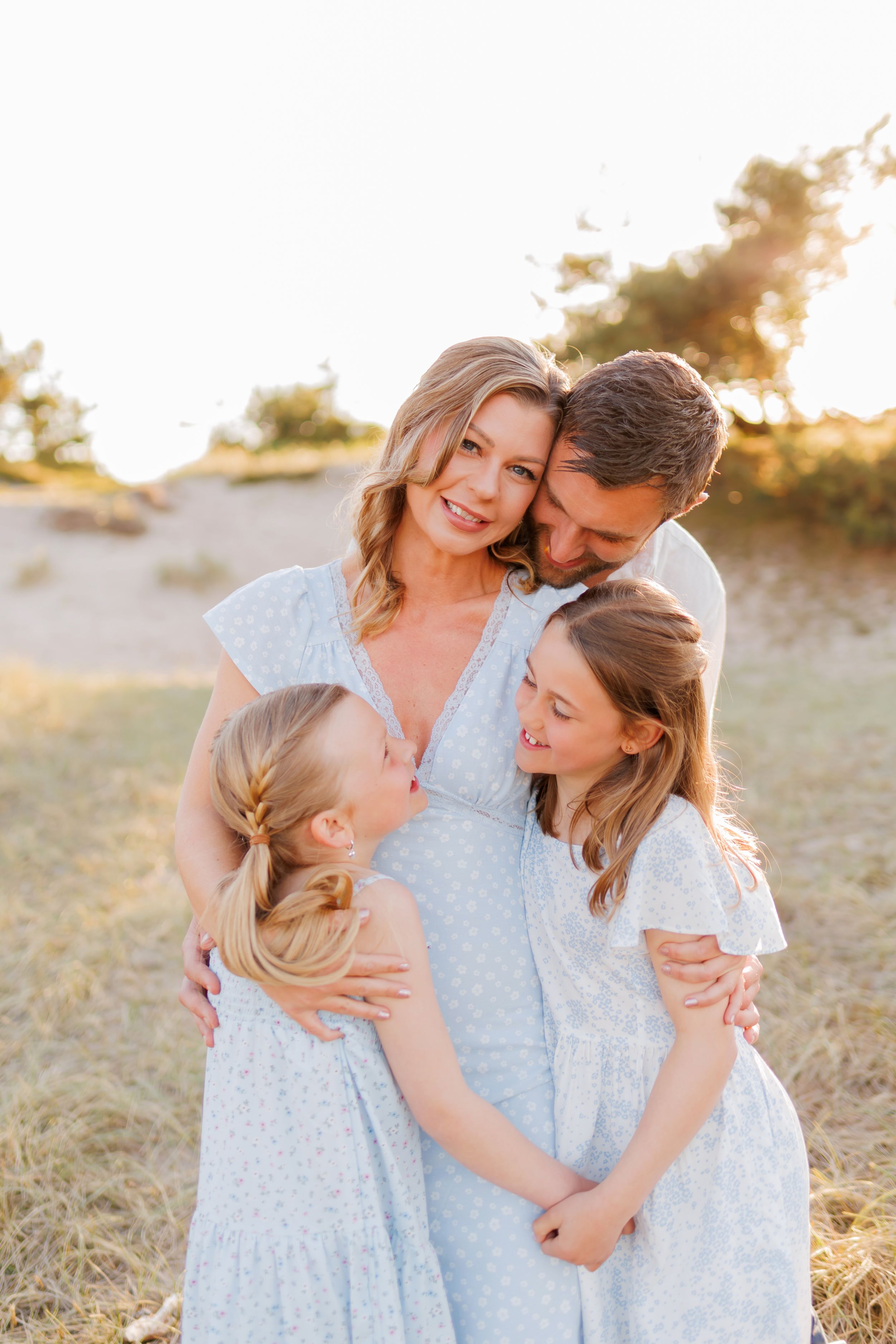 A happy family of four poses in a golden field at sunset, with two blonde girls in light blue dresses hugging their parents.
