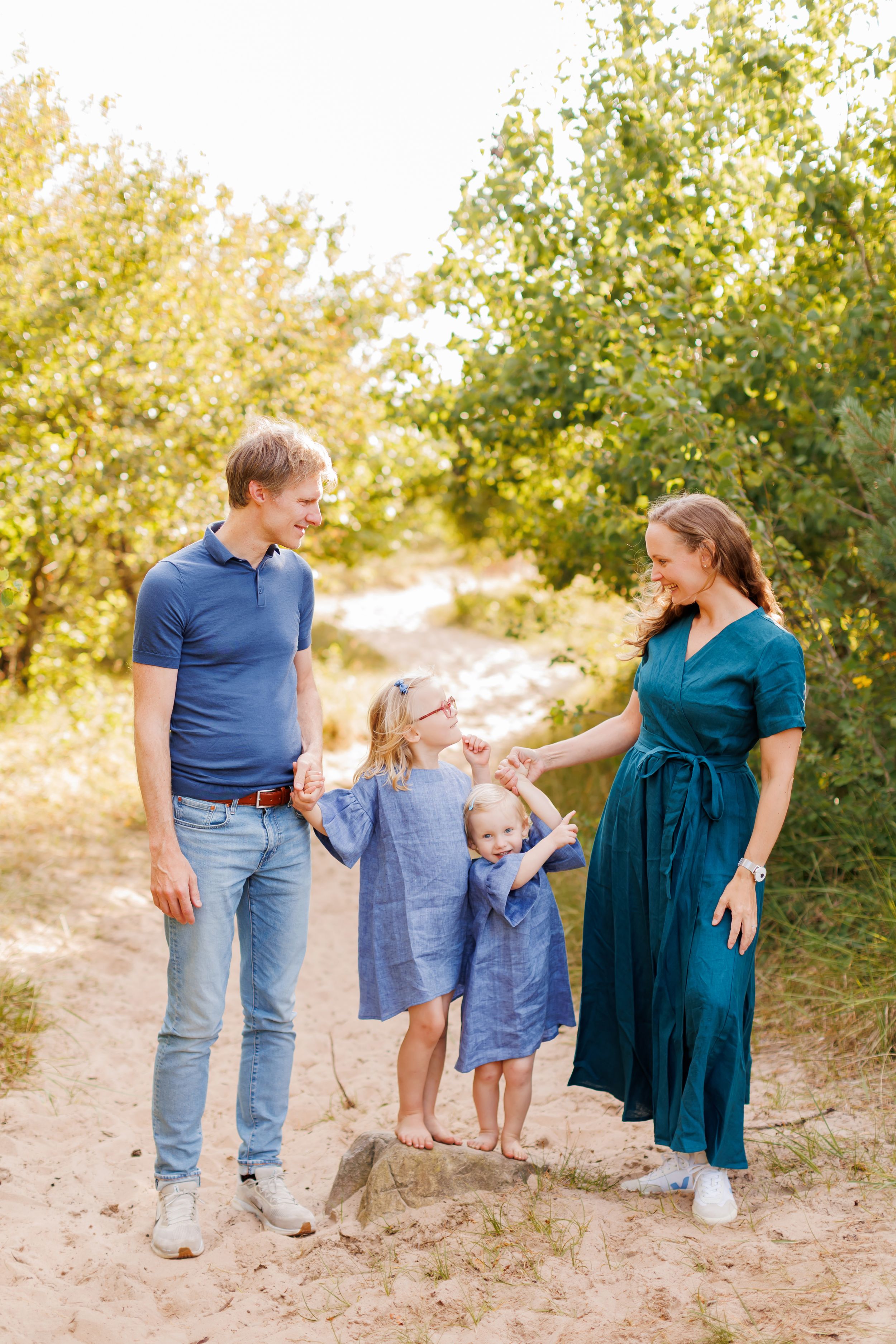 Happy family of four in blue outfits posing on a sandy path surrounded by green trees on a sunny day.
