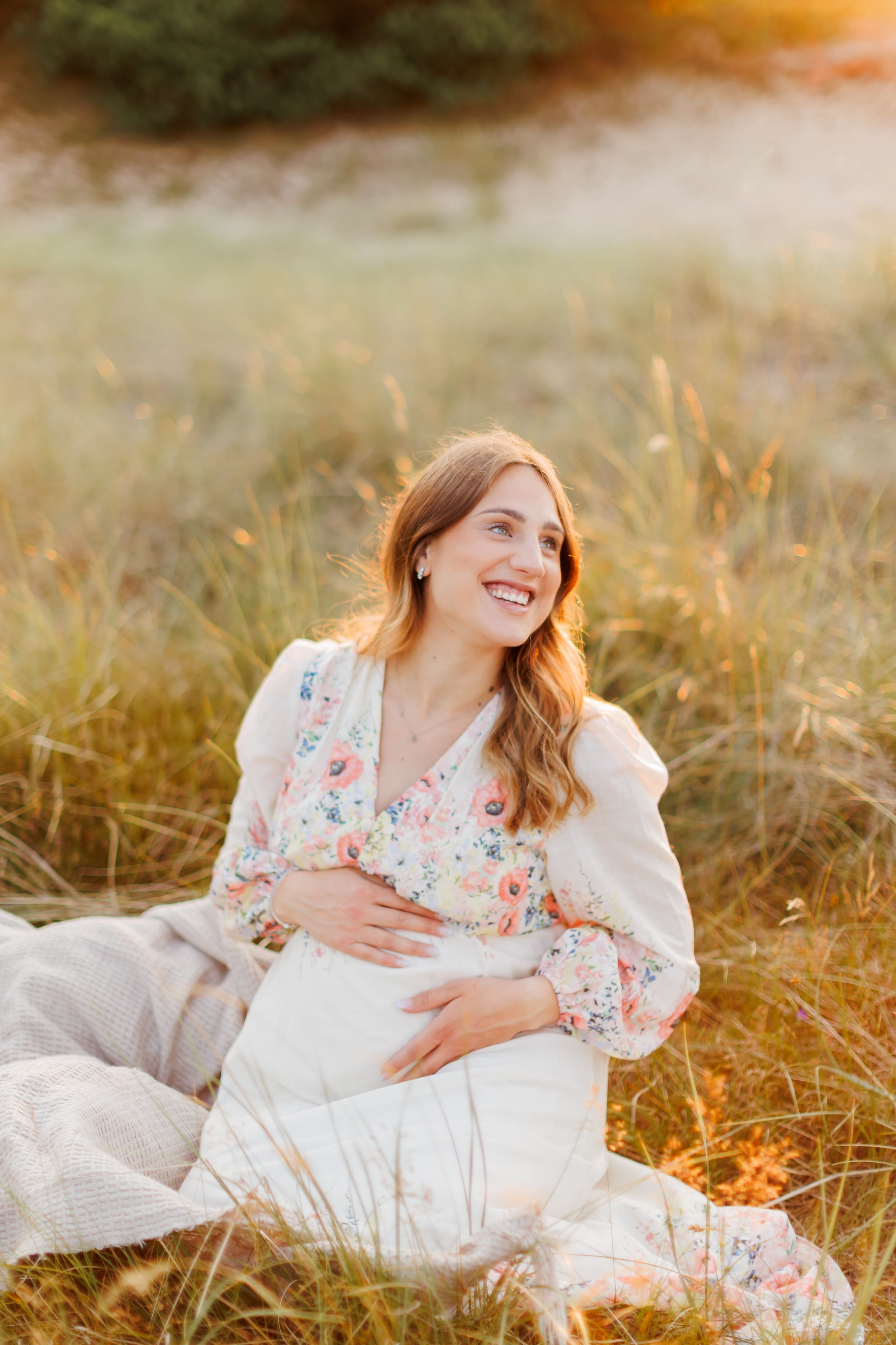 Smiling pregnant woman in floral dress sitting in golden field during sunset maternity photo session.