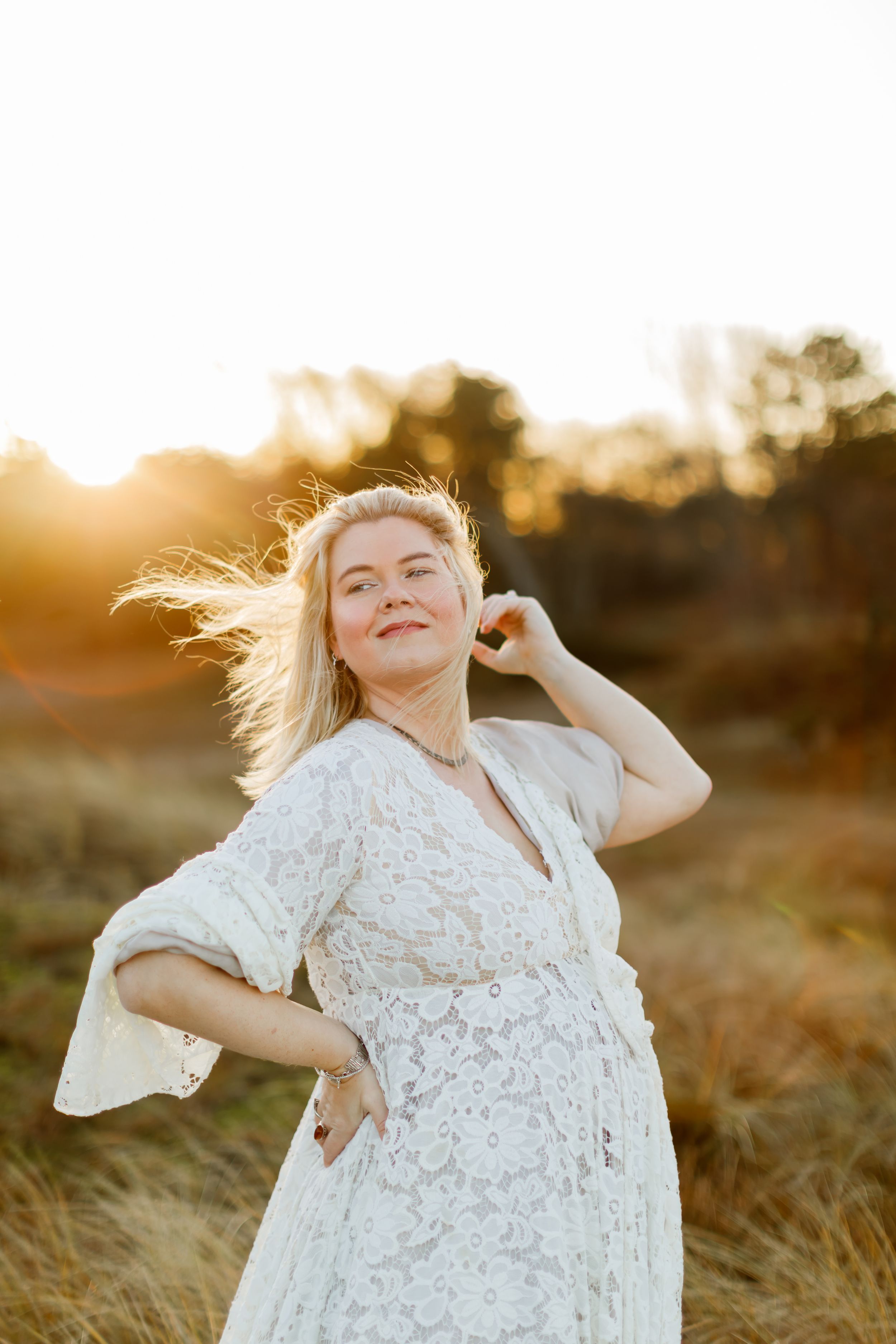 Blonde woman in white lace dress poses in golden field at sunset, hair blowing in the wind.