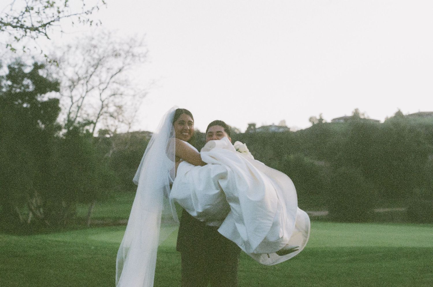 Romantic moment captured as groom carries bride in flowing white dress across green lawn.