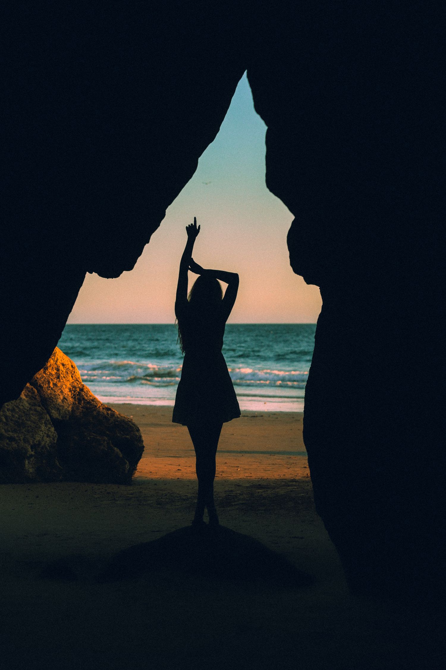 Silhouette of figure with raised arm stands in coastal cave opening framing beach and ocean at sunset.