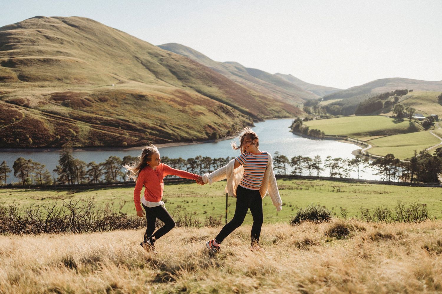 Two people running through a grassy hillside overlooking a scenic lake and mountains in the distance.
