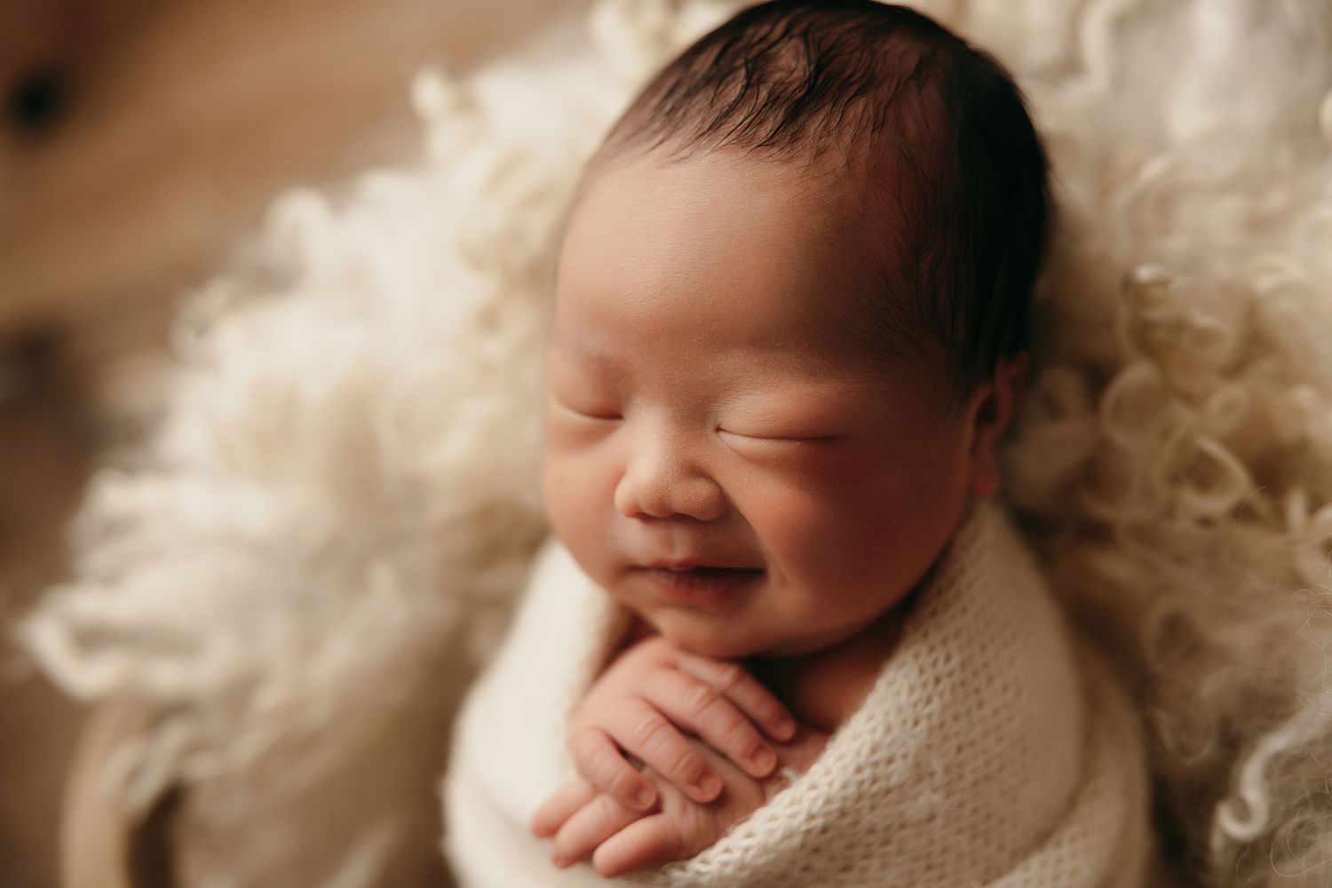 Peacefully sleeping newborn wrapped in soft white blanket with gentle smile on face during professional photoshoot.