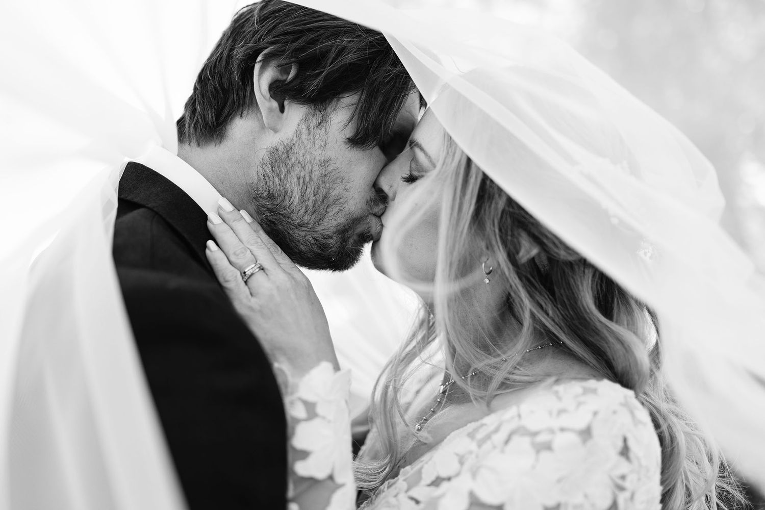 Bride and groom sharing a kiss on a sunlit forest path surrounded by lush green foliage.