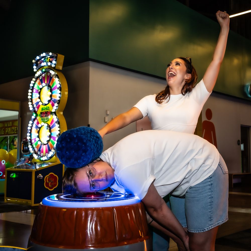 Scripted image of a woman holding a whack-a-mole hammer against another woman's head while raising her arm in triumph