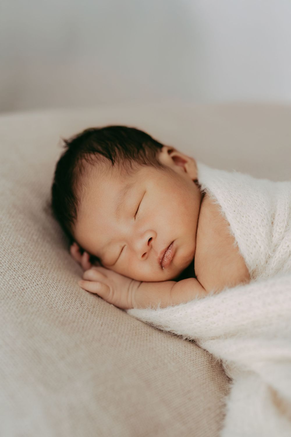 Peaceful sleeping newborn baby wrapped in white blanket on textured neutral background.
