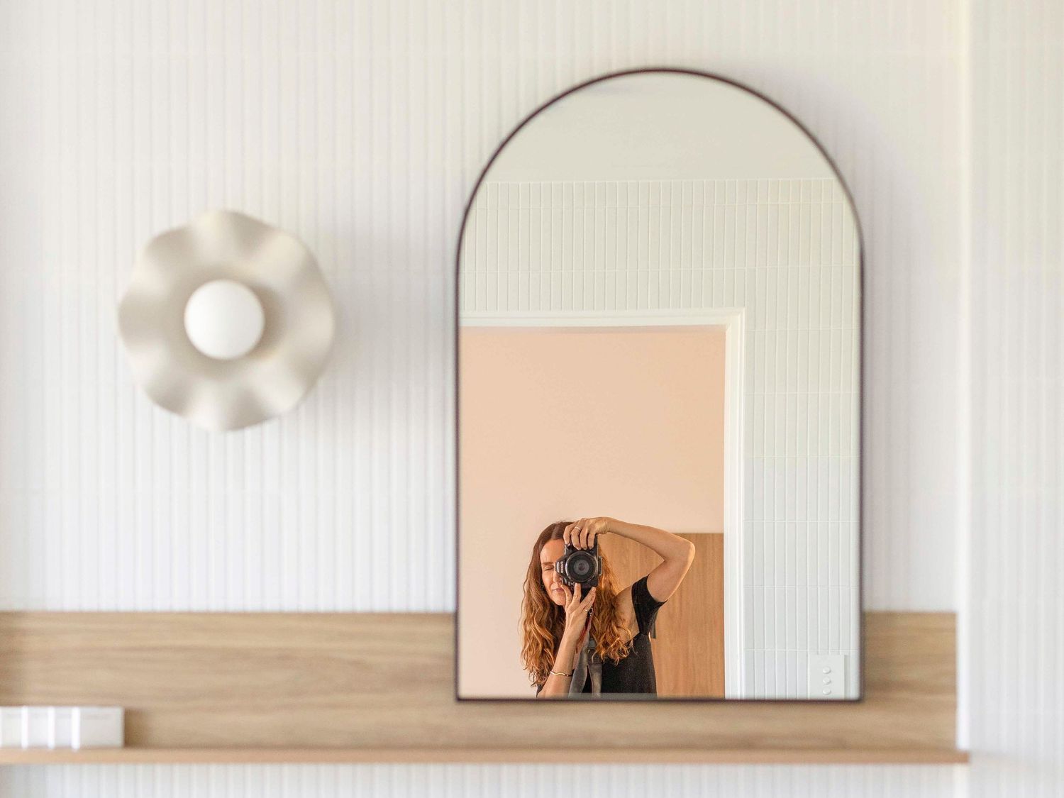 Minimalist arched mirror portrait with floral sconce and decorative artwork on white wall.