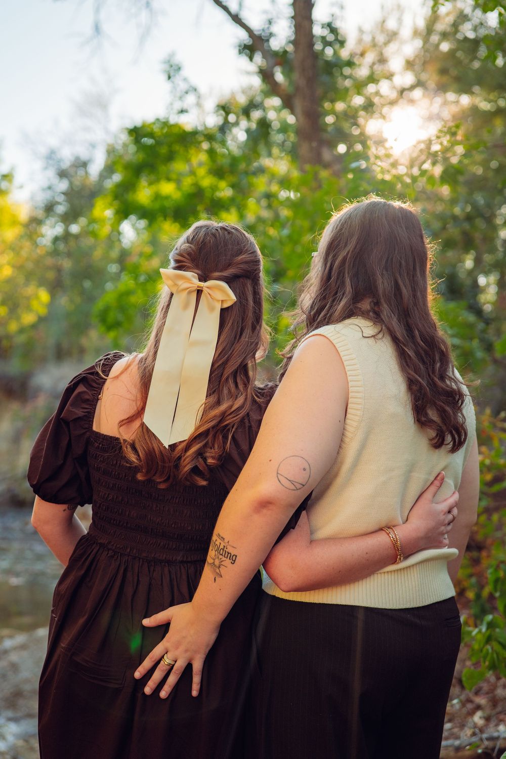 Two people stand close together outdoors with one holding a book and wearing a brown dress.