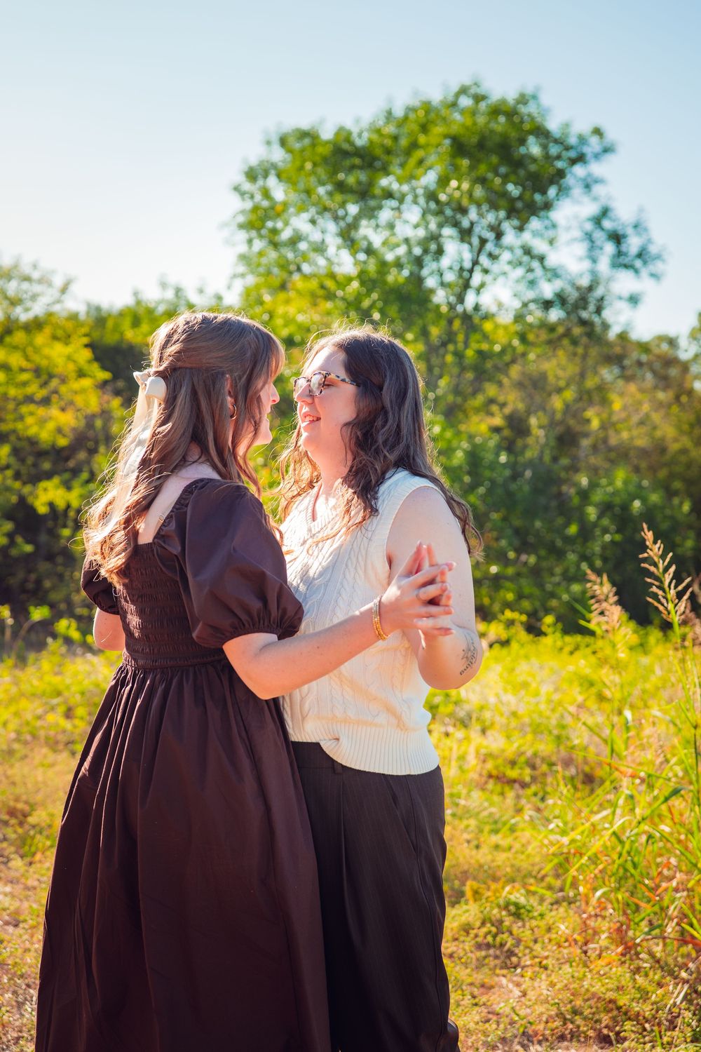 Two people share a joyful embrace in a sunlit meadow with green trees in the background.