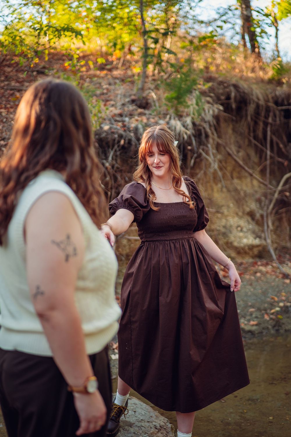 Person in a brown dress twirls playfully by a creek while another watches.