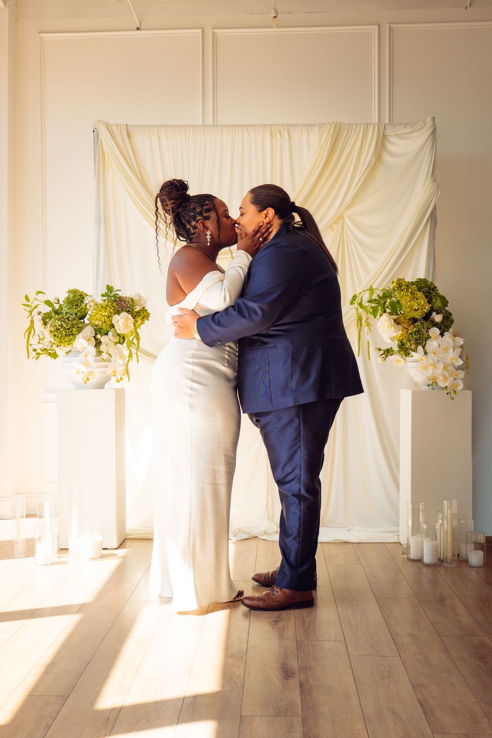 Couple shares first kiss at wedding ceremony with white backdrop and floral arrangements.