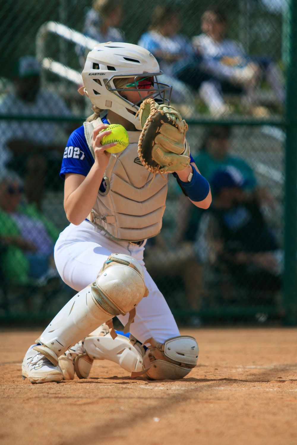 A catcher in protective gear crouches behind home plate with ball in glove during a softball game.