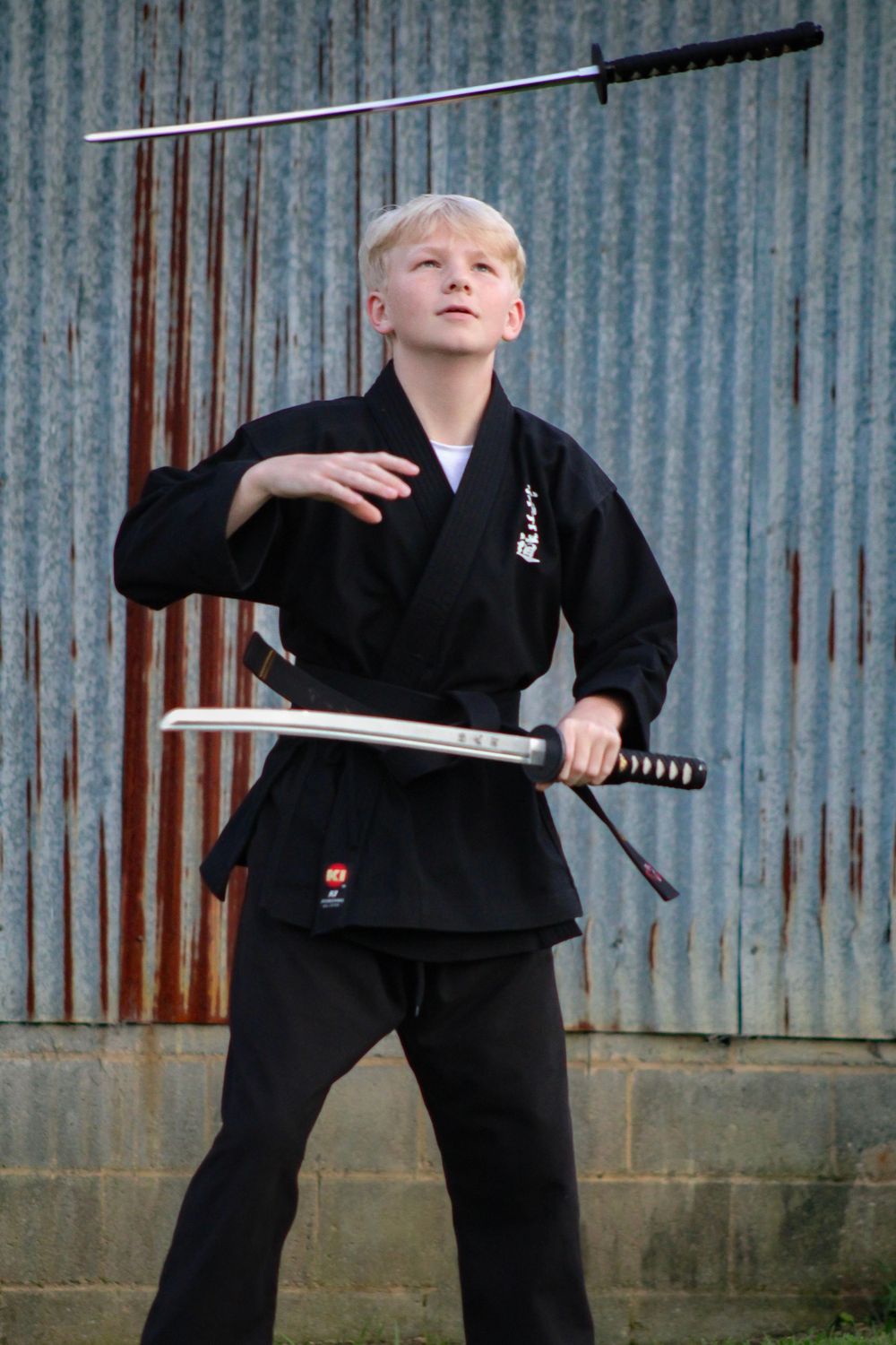 Martial arts practitioner in black uniform executing sword technique against metal wall.
