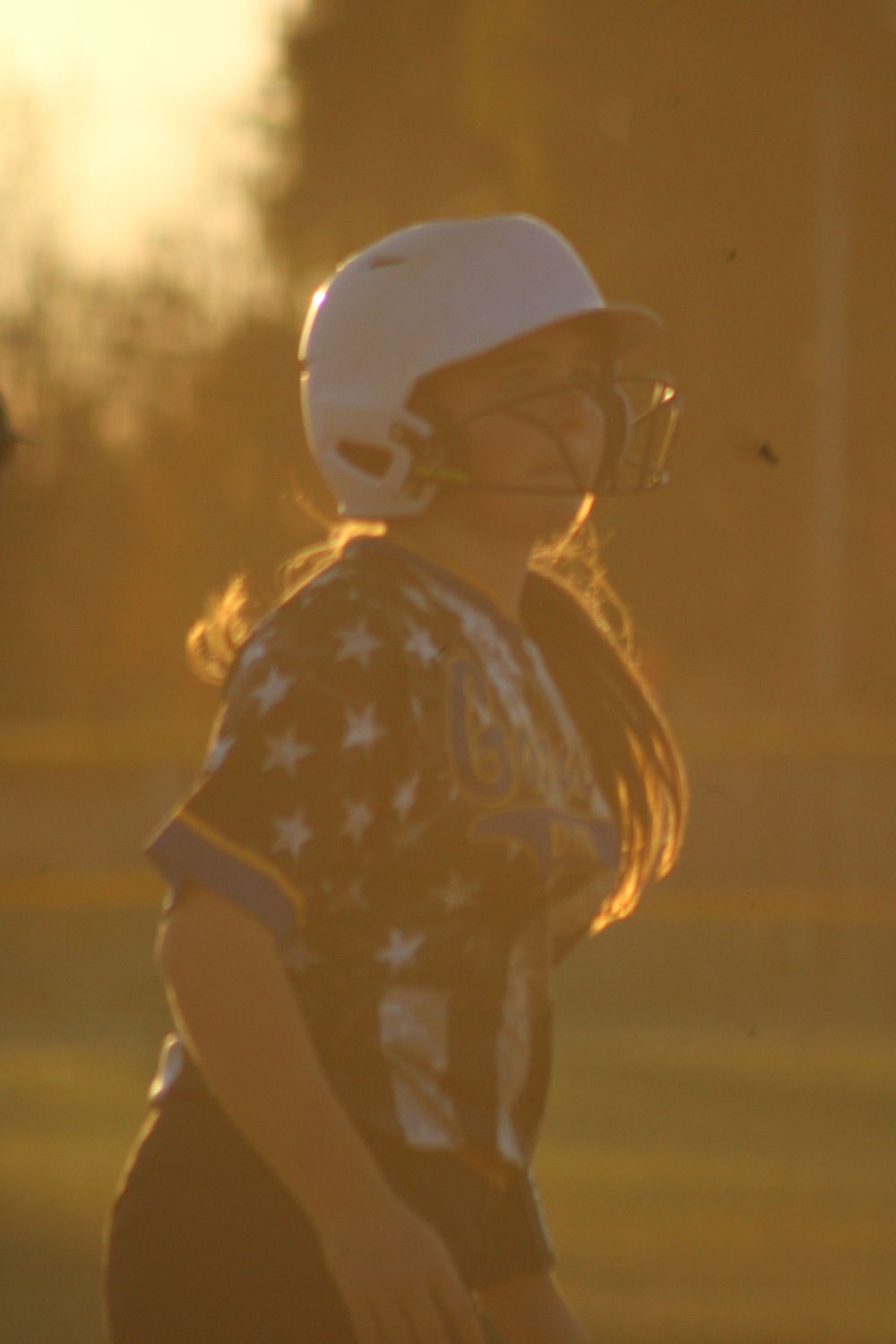 Softball player in stars and stripes uniform stands in golden sunset light.