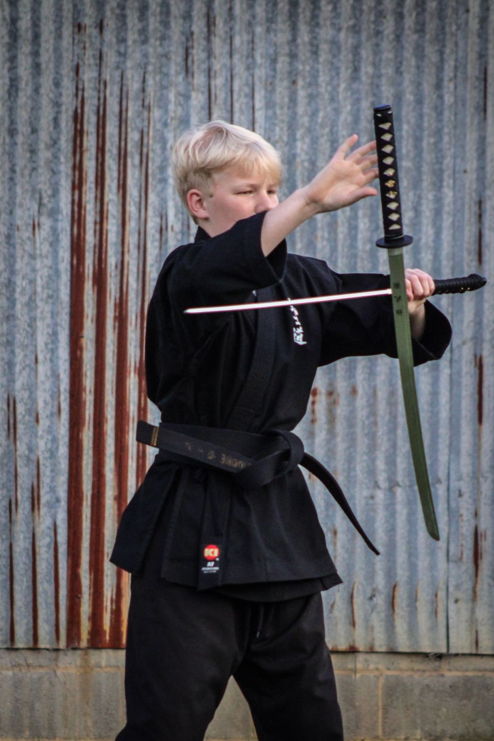 A martial artist in black uniform practicing with a katana sword against a corrugated metal background.